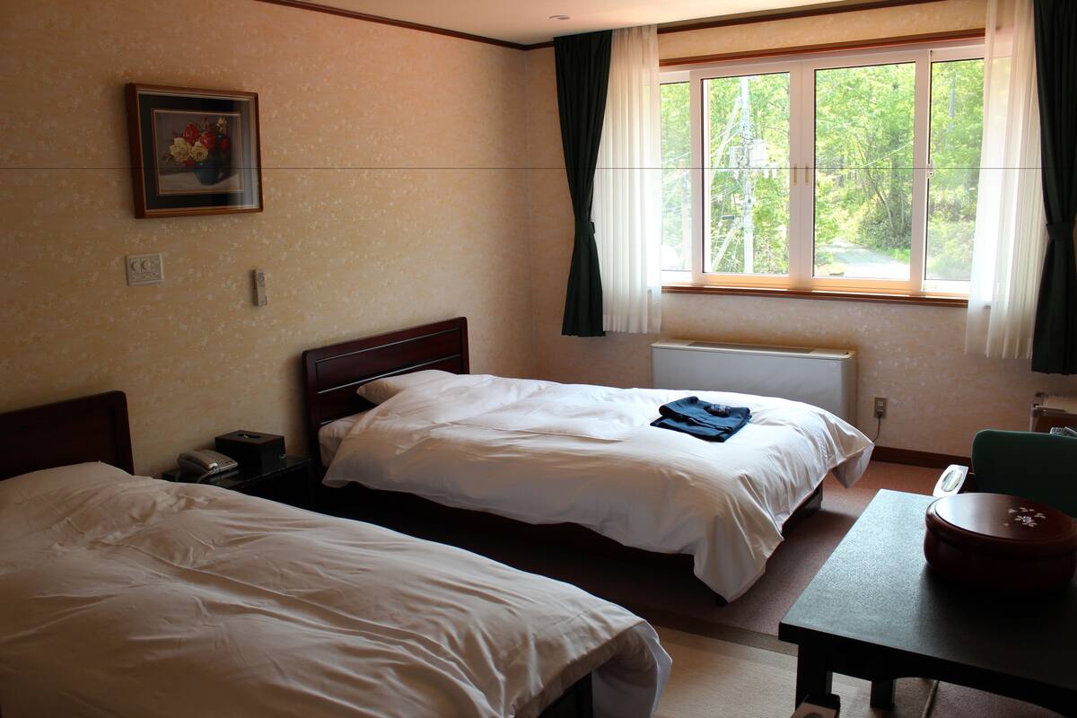 Habitación de hotel con dos camas individuales, una mesita, una silla, un radiador y una ventana con cortinas.