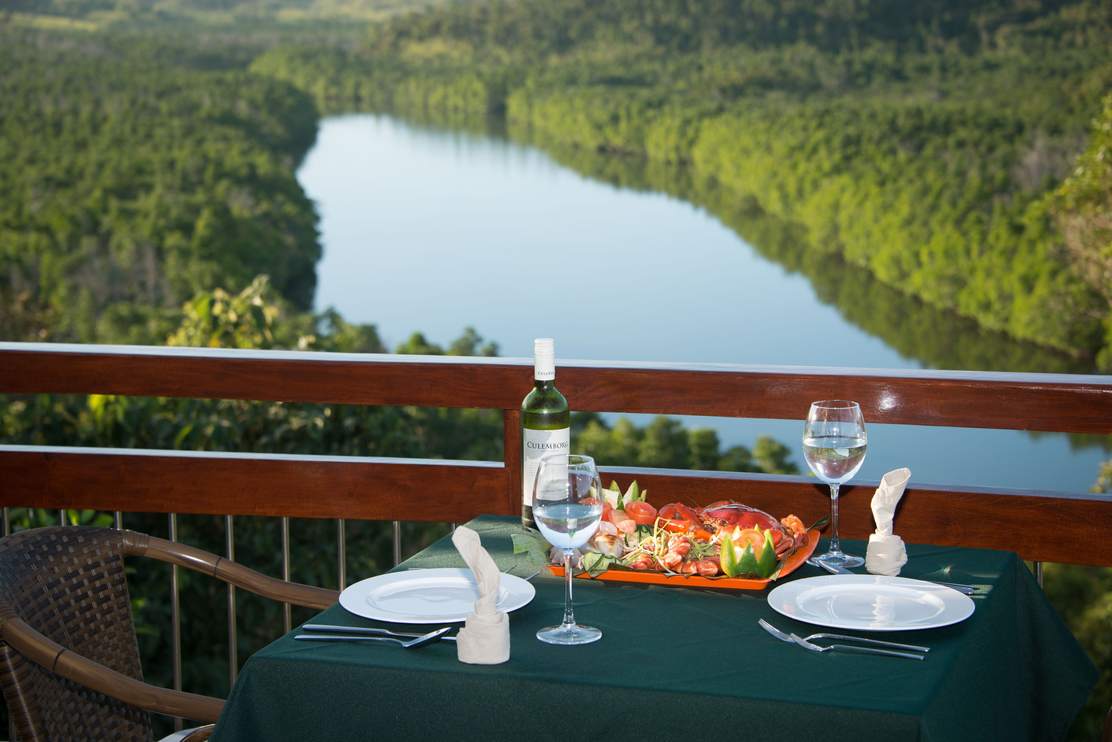 A table set for two with a bottle of wine, glasses, and a plate of food, overlooking a lake and forest.