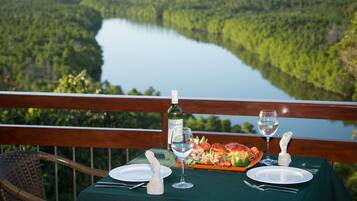 A table set for two with a bottle of wine, glasses, and a plate of food, overlooking a lake and forest.