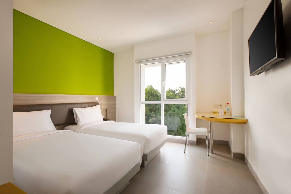 Een hotelkamer met twee bedden, een groene accentmuur, een raam met uitzicht, een klein bureau en een aan de muur bevestigde televisie.