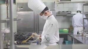 Un chef, vêtu d’un uniforme blanc et coiffé d’un chapeau, cuisine dans une cuisine en acier inoxydable équipée d’une grande cuisinière et de divers ustensiles de cuisine.