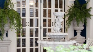 Una fontana in pietra a più livelli con acqua che scorre, circondata da colonne bianche e felci in vaso, con sullo sfondo una porta bianca con pannelli in vetro.