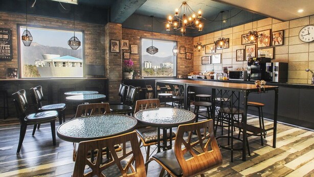 Un café chaleureux avec des tables et des chaises rondes, un coin bar et une grande fenêtre donnant sur les bâtiments.