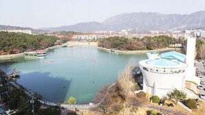 Une vaste étendue d’eau où naviguent des bateaux, un bâtiment blanc surmonté d’un toit bleu, ainsi que des arbres et des montagnes environnants.
