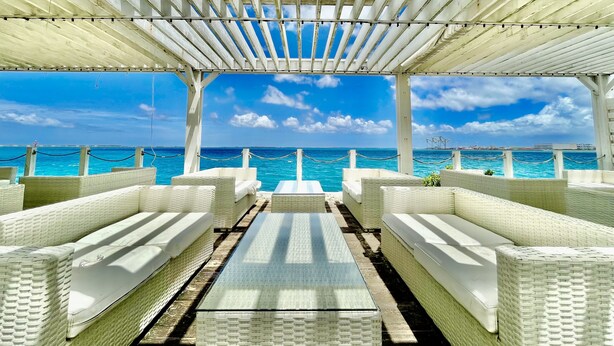 A covered outdoor seating area with white wicker furniture and a view of the ocean. - Hotel Umizola (Naha)