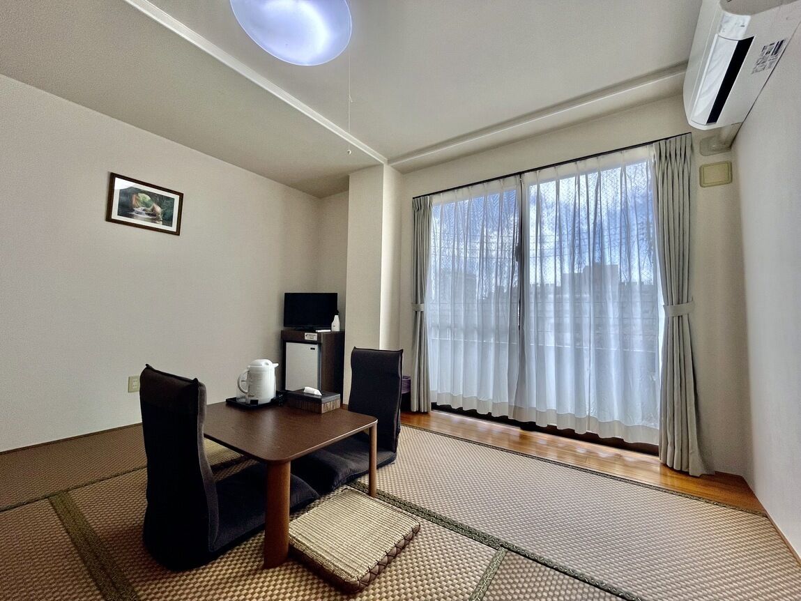 Una habitación con una mesa de comedor, sillas, una televisión, un cuadro en la pared y un gran ventanal con cortinas.