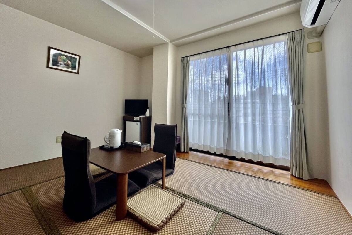 A room with a dining table, chairs, a television, a picture on the wall, and a large window with curtains.