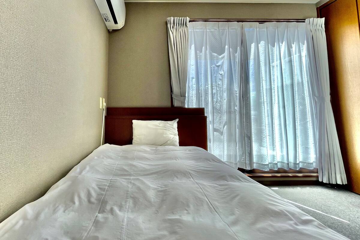 A single bed with a wooden headboard, a white pillow, and a plain wall with a textured surface.