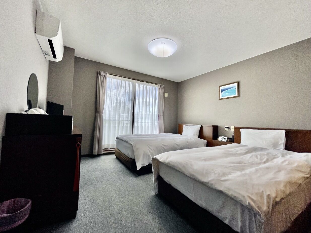 Habitación de hotel con dos camas, un escritorio, una silla, un espejo y un ventanal con cortinas.