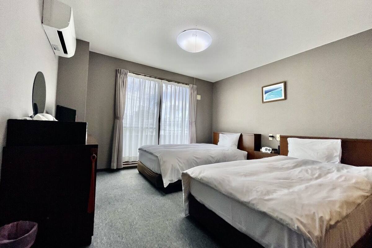 A hotel room with two beds, a desk, a chair, a mirror, and a window with curtains.