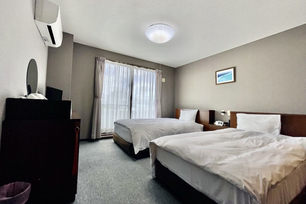 Quarto de hotel com duas camas, uma escrivaninha, uma cadeira, um espelho e uma janela com cortinas.