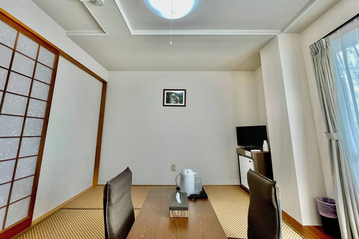 A hotel room with tatami mat flooring, a wooden dining table, a television, and a small kitchenette.