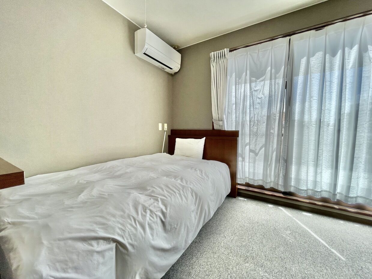 Habitación de hotel con cama, aire acondicionado, ventana con cortinas translúcidas y cabecero de madera.