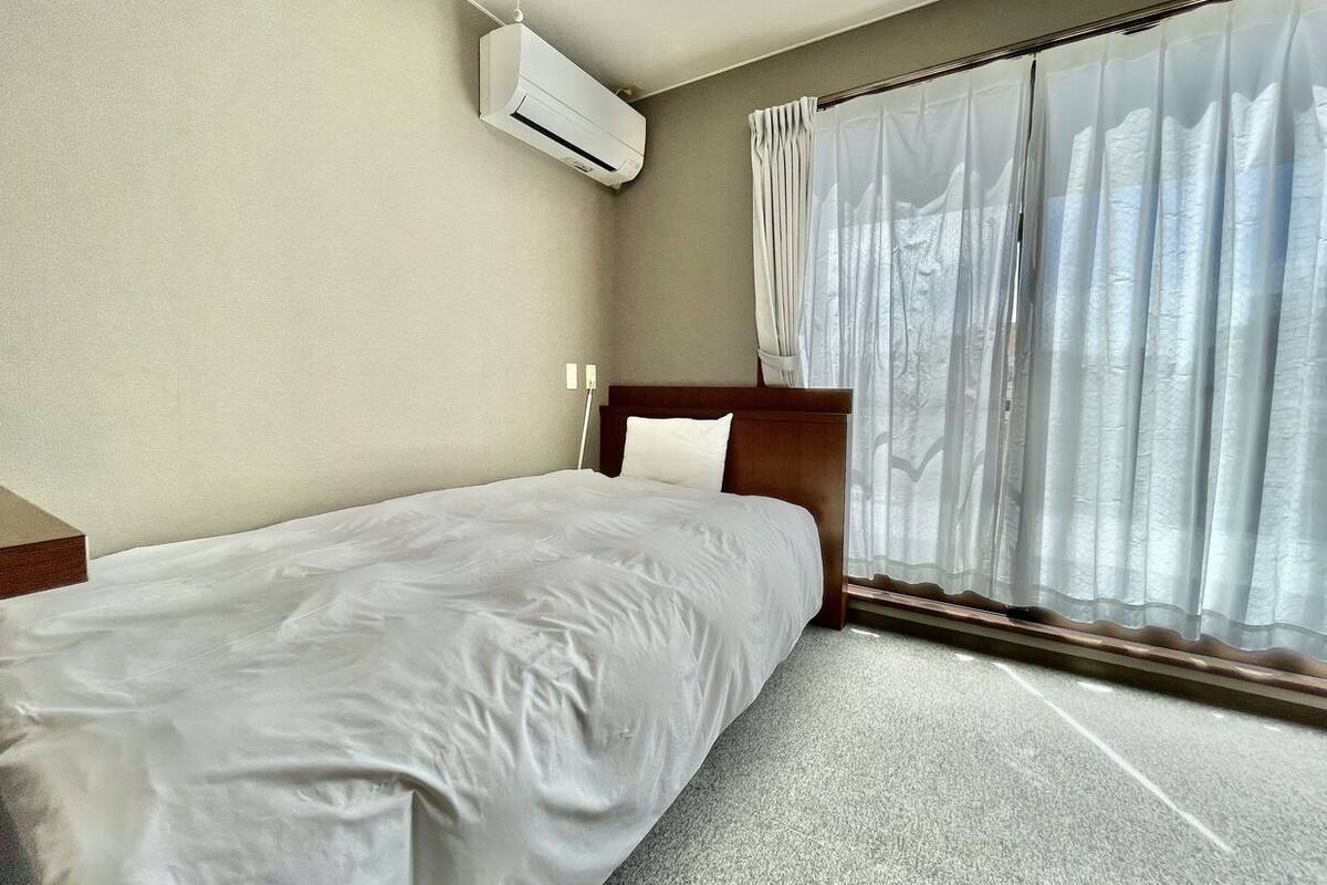 A hotel room with a bed, an air conditioning unit, a window with sheer curtains, and a wooden headboard.