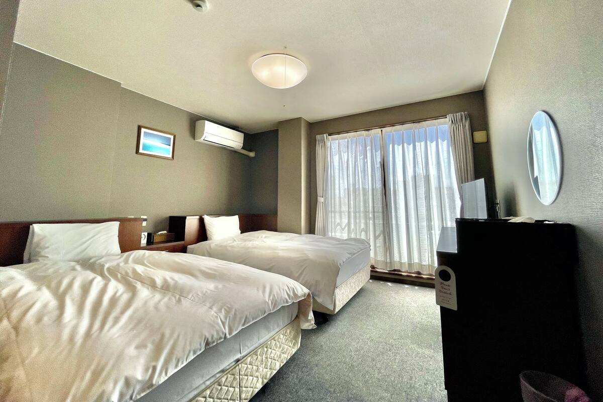 A hotel room with two beds, a flat-screen TV, a mirror, and a window with curtains.