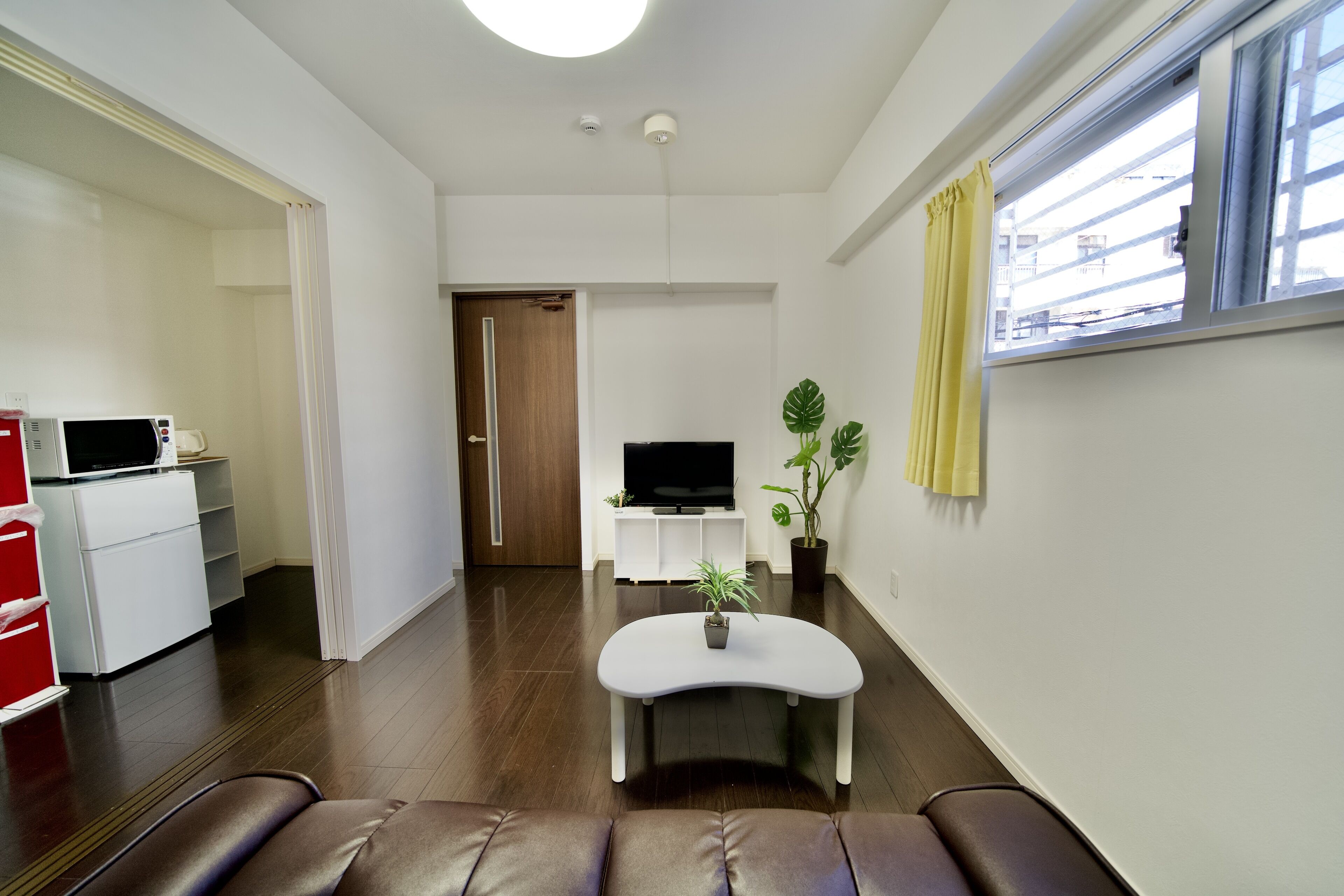 Een moderne woonkamer met een bruine leren bank, een witte salontafel, een flatscreen televisie, een kleine potplant en een raam met gele gordijnen.