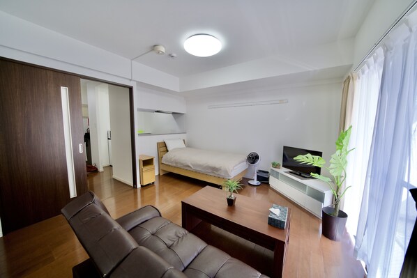 A modern, well-lit room with a bed, a desk, a chair, a TV, and a potted plant. - Villa Izumizaki (Naha)