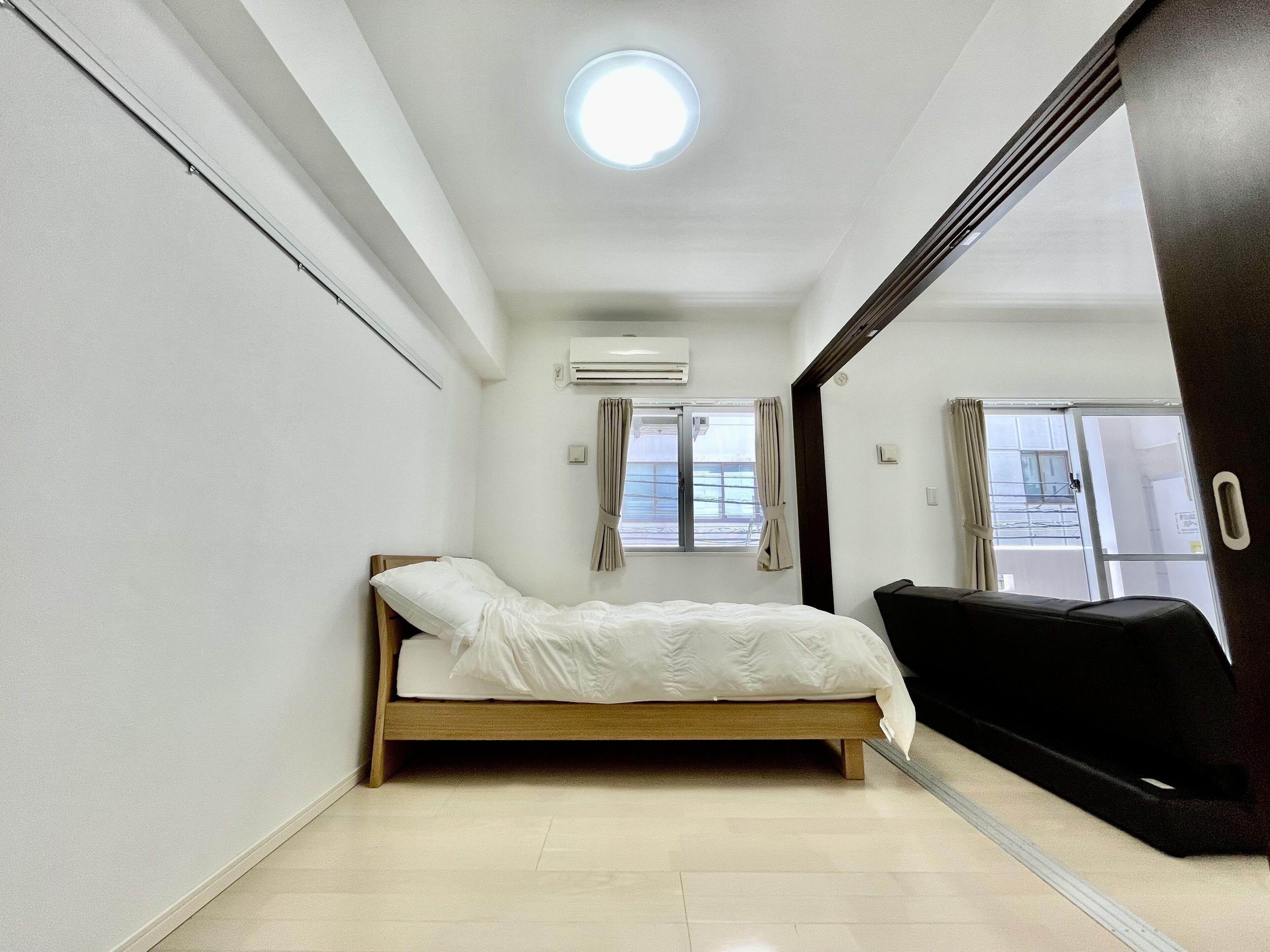 A small, clean room with a single bed, a window with curtains, an air conditioning unit, and a door leading to another room.