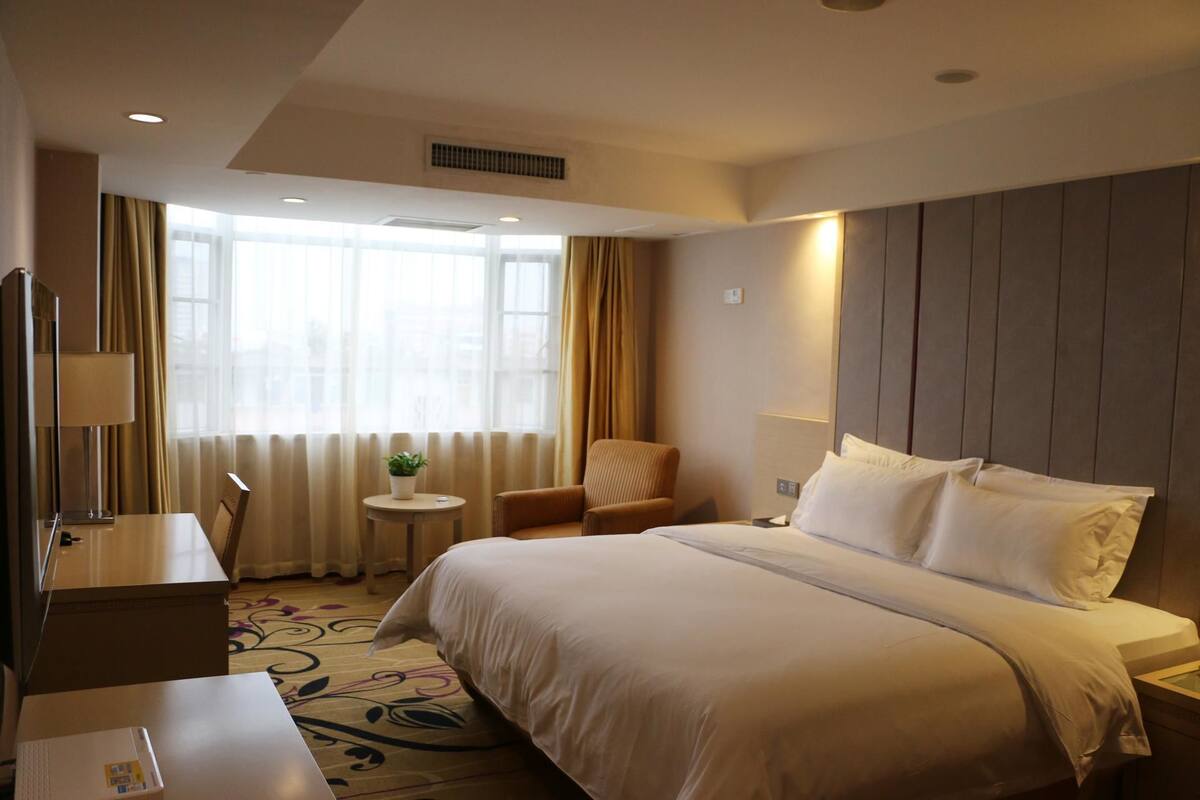 Ein Hotelzimmer mit einem großen Bett, einem Schreibtisch, einem Sessel, einem kleinen Tisch mit Pflanze und einem Vorhangfenster.