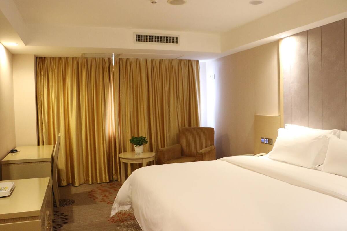Ein Hotelzimmer mit einem Bett, einem Schreibtisch, einem Stuhl, einem kleinen Tisch mit Pflanze und einer an der Wand befestigten Bedieneinheit.