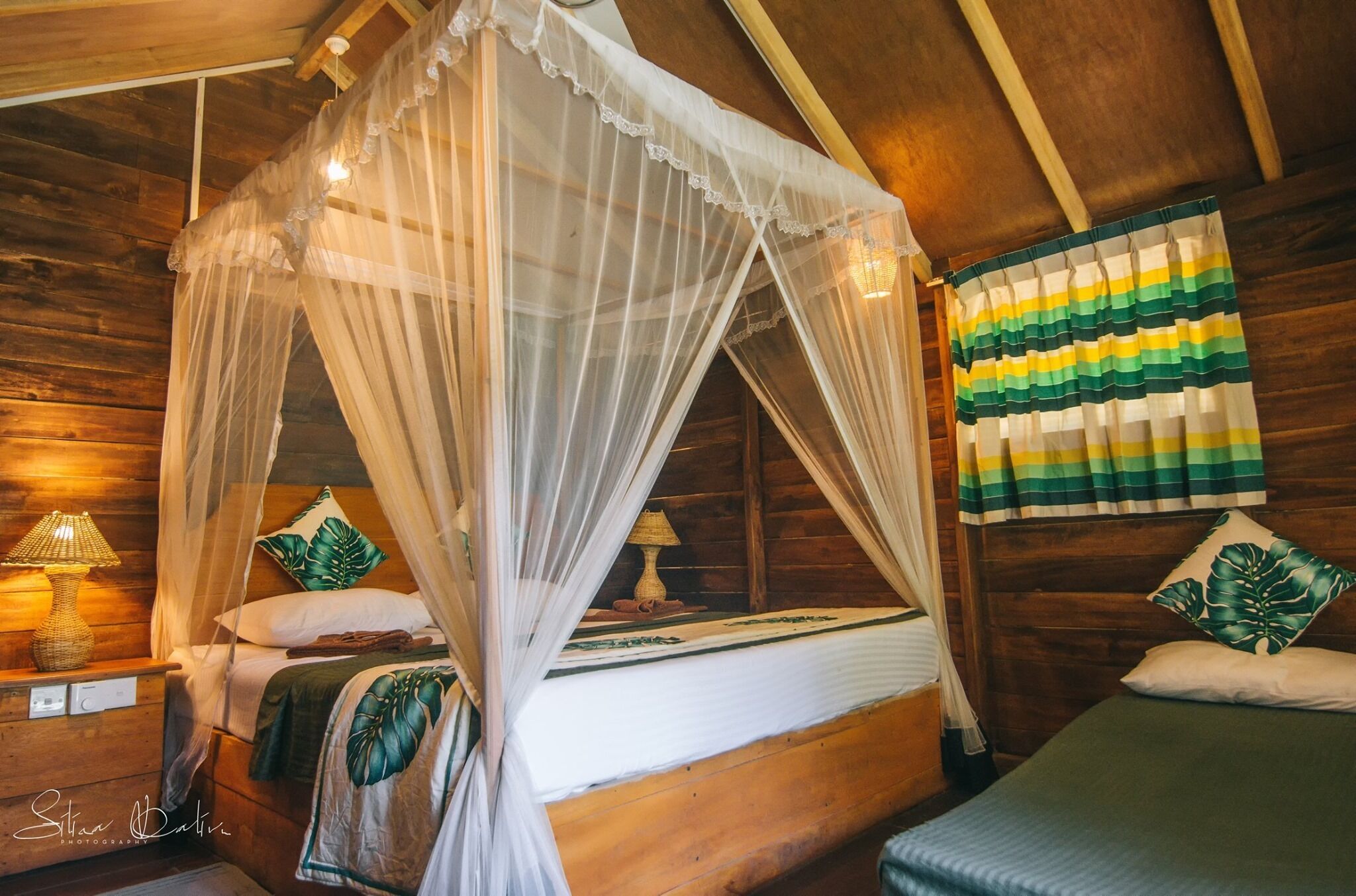 Een slaapkamer in een houten blokhut met een hemelbed, een bedlamp en kussens met een tropisch thema.