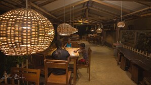 A rustic dining area with wooden tables and chairs, large woven light fixtures, and a stone wall in the background. - 360 Rainforest (Deniyaya)