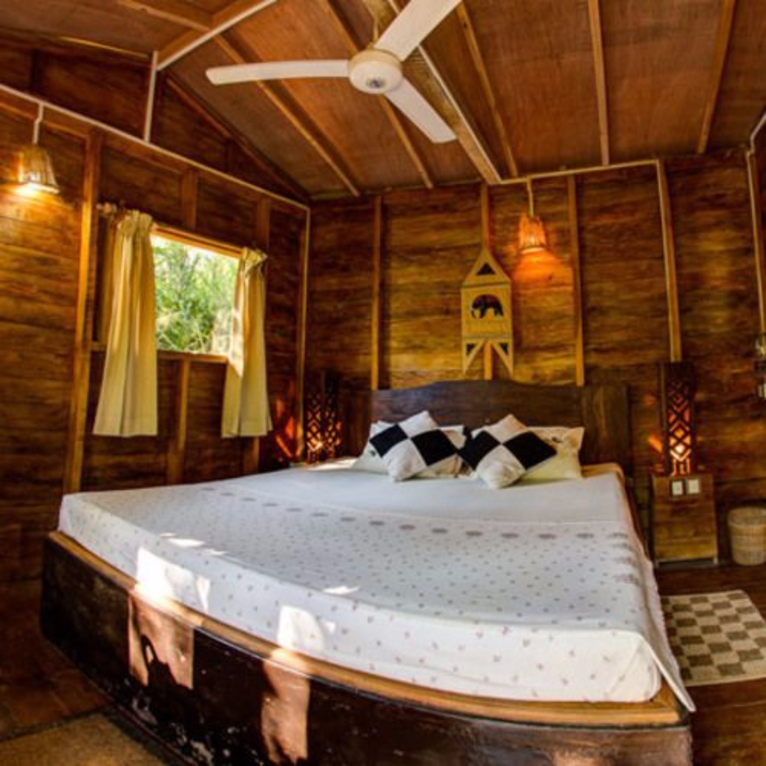 A wooden cabin bedroom with a bed, a ceiling fan, a window with curtains, and a birdhouse decoration.