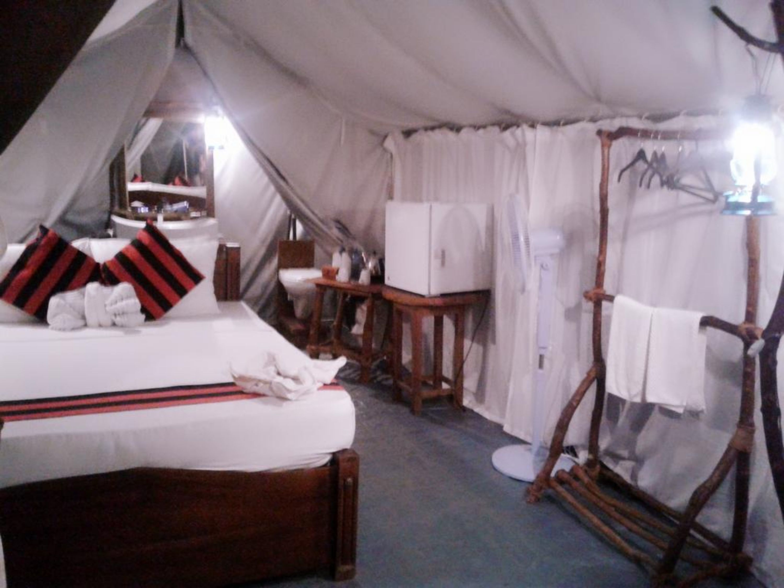 Una habitación tipo tienda de campaña con una cama, una mesita, una silla, un espejo y un tendedero.