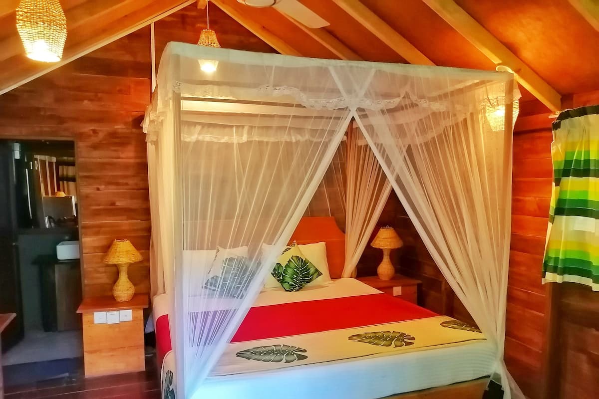 A wooden room with a four-poster bed, a red bedspread, and a canopy. There are two lamps on wooden nightstands.