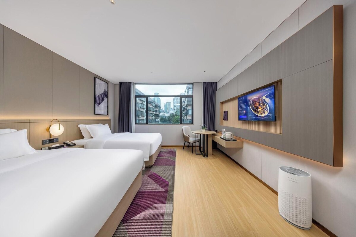 A hotel room with two beds, a desk, a chair, a TV, and a large window with a city view.