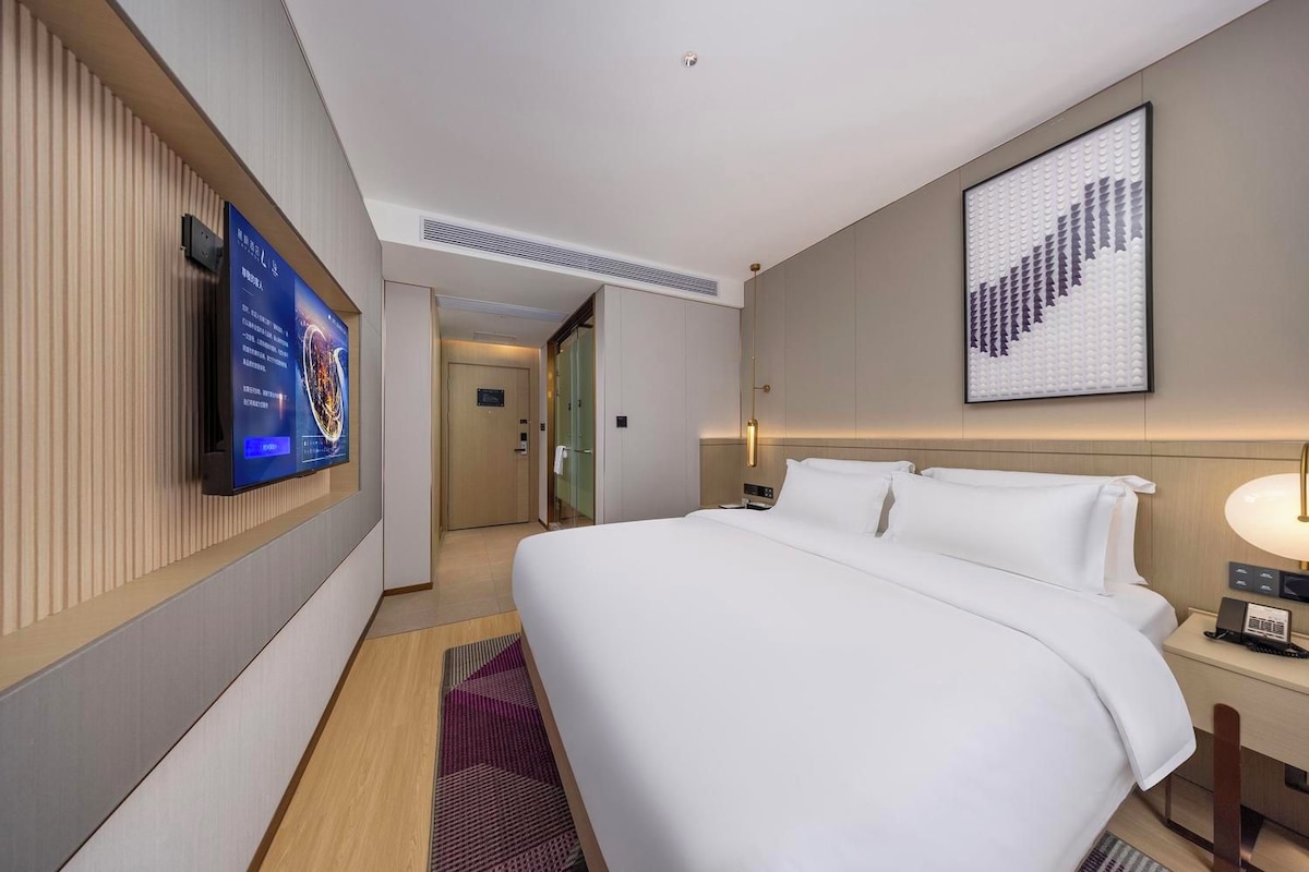A hotel room with a large bed, a flat-screen TV mounted on the wall, a bedside table with a lamp, and a framed artwork above the bed.