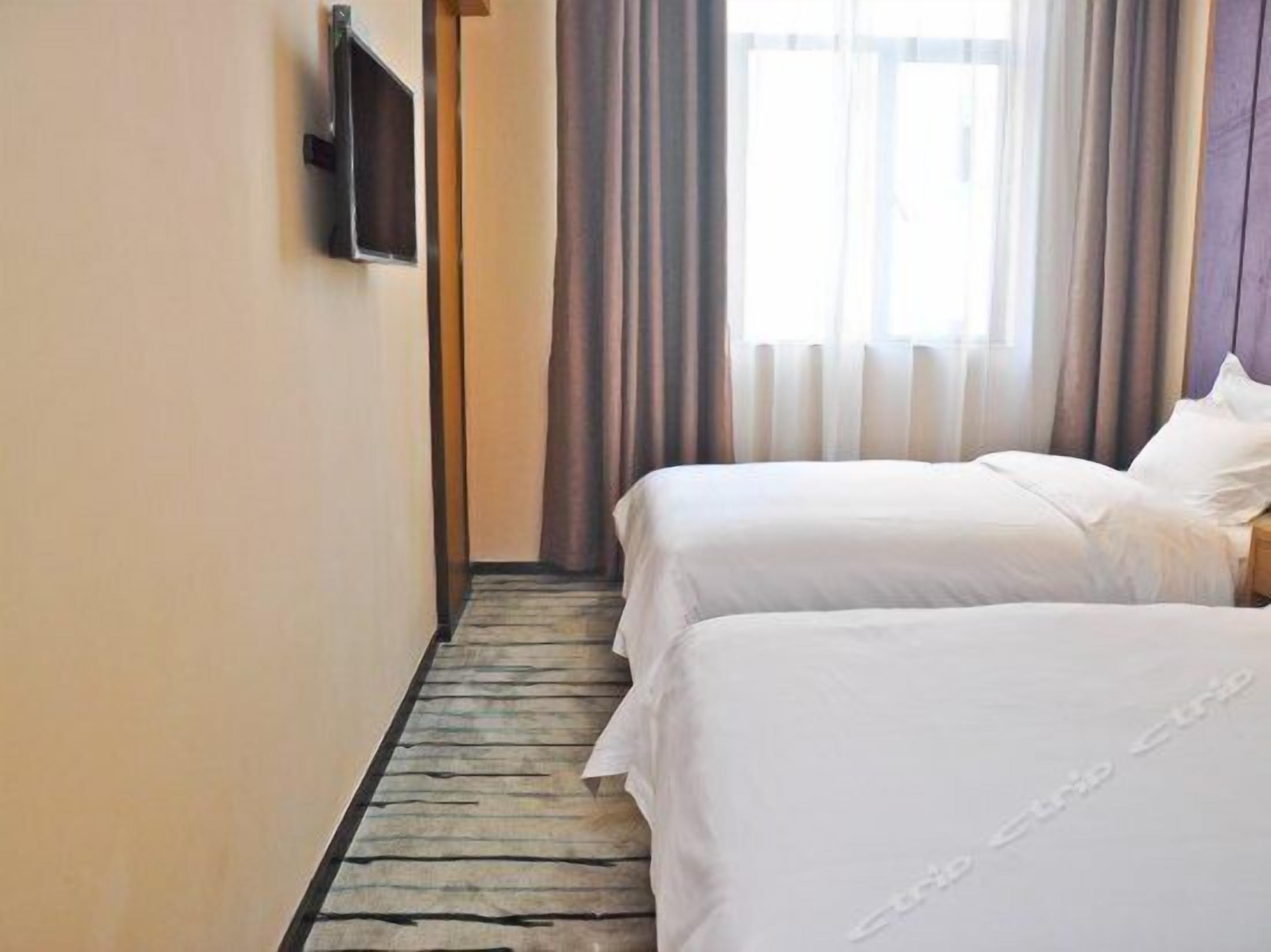 Een hotelkamer met twee bedden, een houten vloer, een aan de muur bevestigde televisie en een raam met gordijnen.