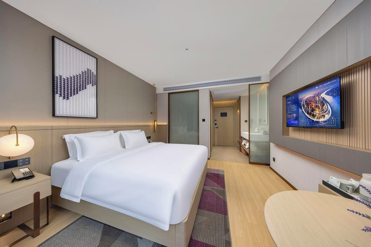 A hotel room with a bed, bedside table, lamp, TV, and a view of the hallway.