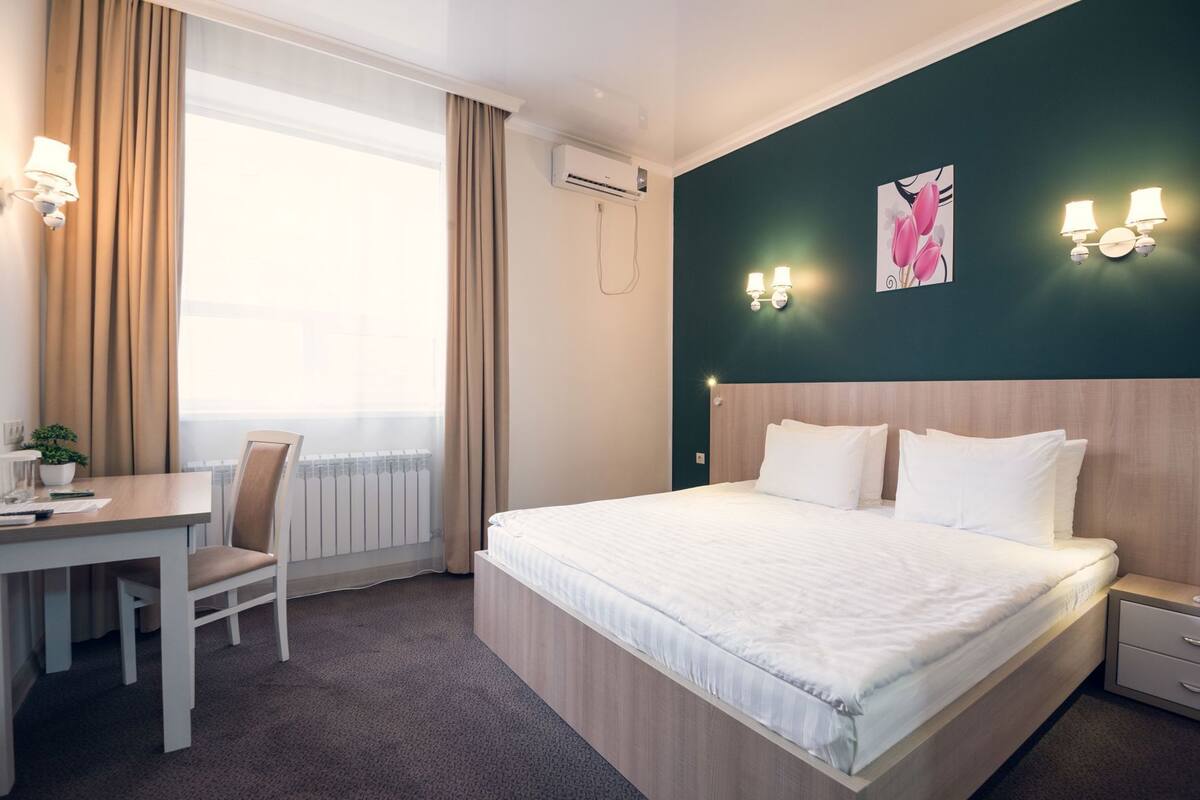 Habitación de hotel con cama, mesita de noche, escritorio, silla, equipo de aire acondicionado y ventana con cortinas.