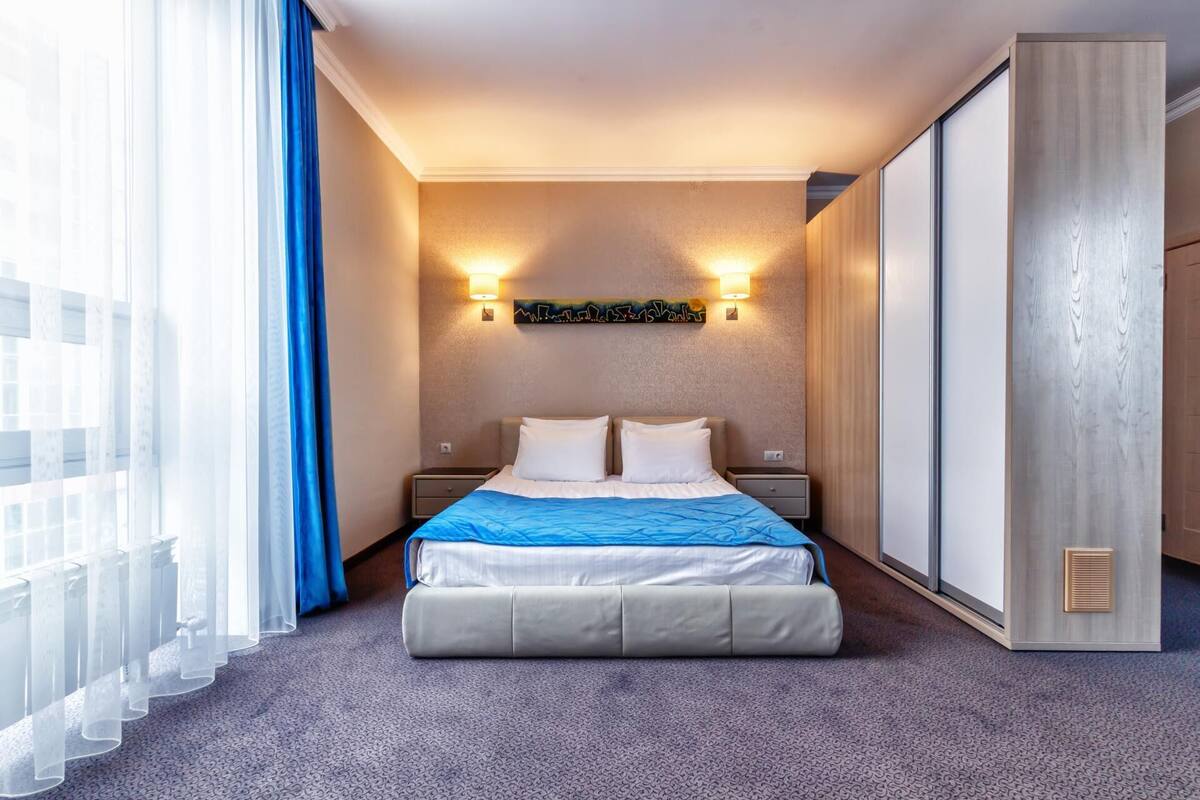 Habitación de hotel con cama, dos mesitas de noche, una lámpara de pared y un ventanal con cortinas.