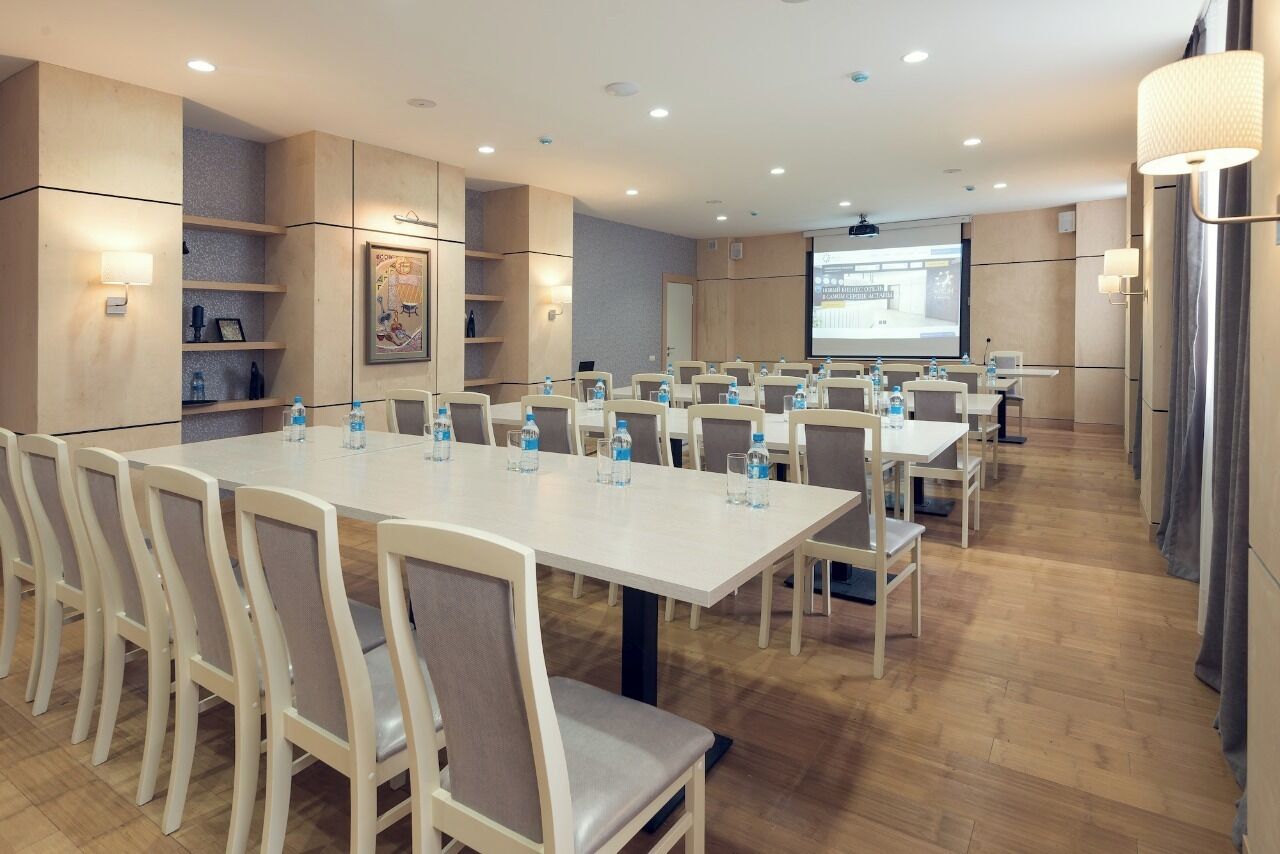 Un salón de conferencias con varias mesas y sillas, equipado con botellas de agua y una pantalla de proyector.