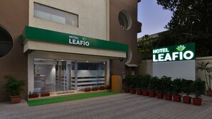 Entrada de hotel com placa verde, portas de vidro e plantas em vasos.