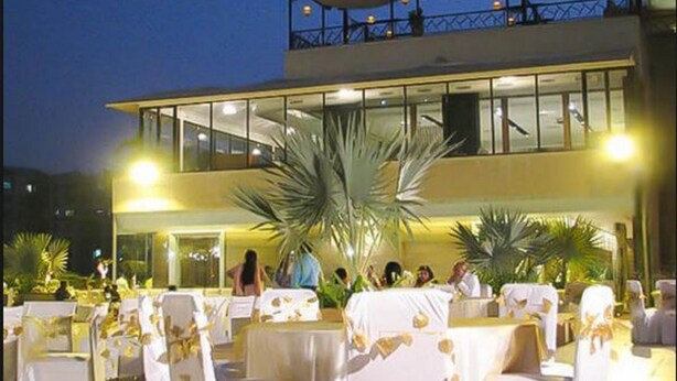 Um espaço para eventos ao ar livre com mesas montadas para convidados, uma grande palmeira e um edifício com varanda ao fundo.