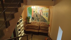 Una escalera conduce a una sala de estar con un sofá de cuero marrón, un estante blanco y vistas a la calle a través de una ventana.