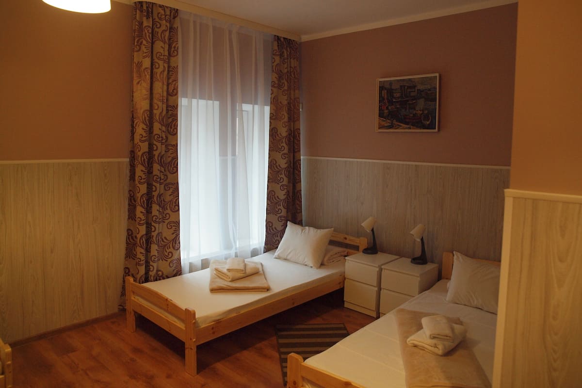 A small, cozy room with two single beds, a wooden floor, a window with curtains, a bedside table with lamps, and a painting on the wall.