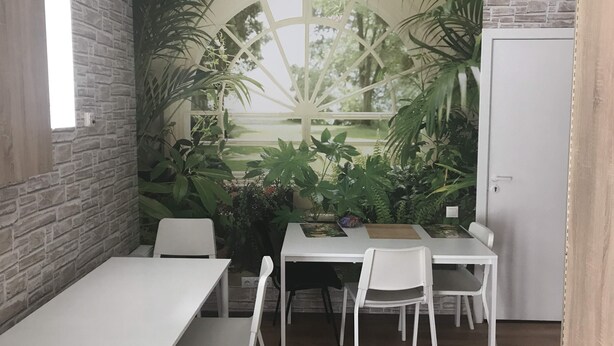 Una habitación con pared de ladrillo, una mesa blanca y dos sillas blancas. Se ve vegetación por la ventana.
