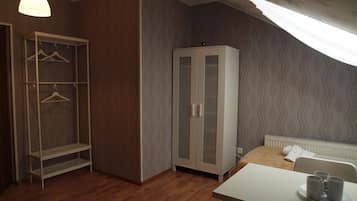 A bedroom with a wooden floor, a white wardrobe, a bedside table with a tray of mugs, and a window with a sheer curtain.