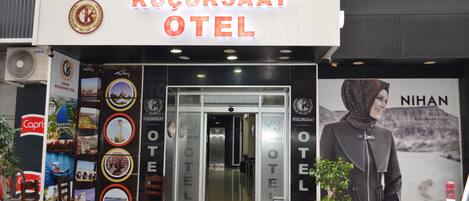 La entrada del Hotel Küçüksaat con un puerta de vidrio, un cartel grande sobre la entrada y un cartel de una persona a la derecha.