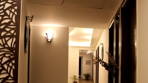 A hotel hallway with a wooden floor, wall-mounted light fixtures, potted plants, and doors leading to rooms.