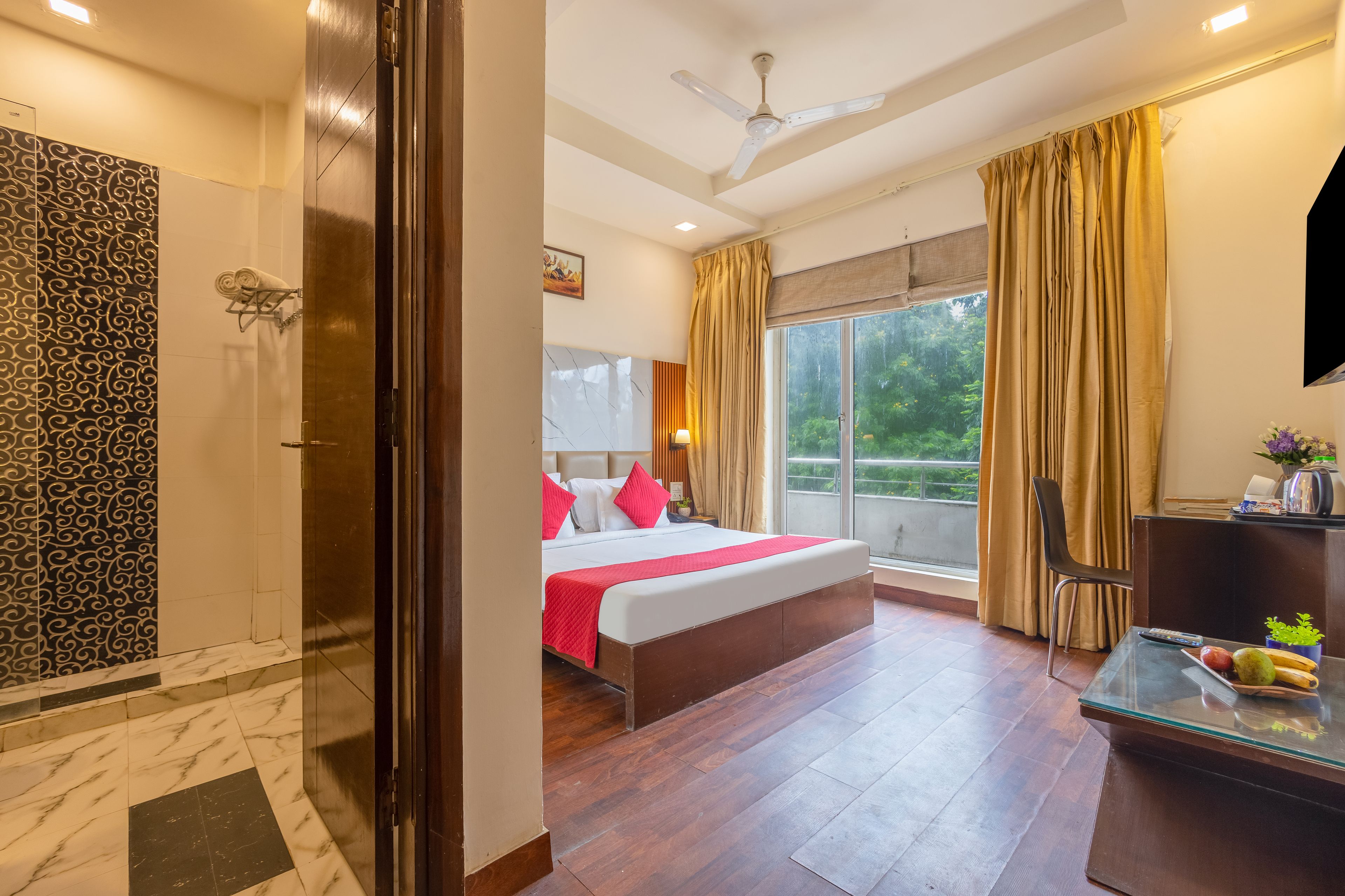 A hotel room with a bed, a desk, a chair, a window with curtains, a ceiling fan, and a shower area.