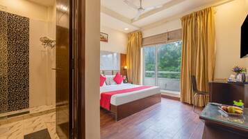 A hotel room with a bed, a desk, a chair, a window with curtains, a ceiling fan, and a shower area.