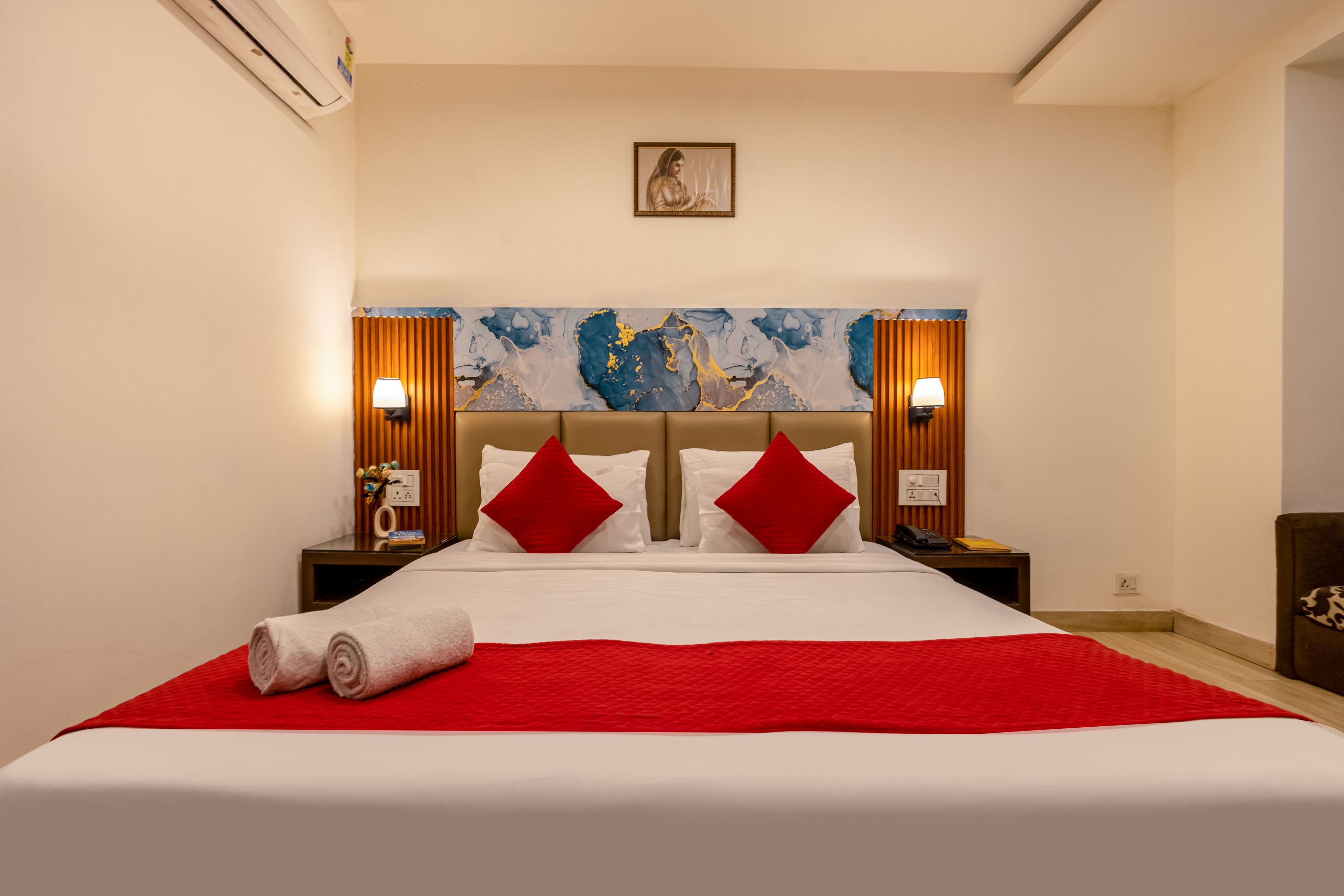 A hotel room with a large bed, red and white bedding, two nightstands with lamps, a painting on the wall, and a red throw blanket.