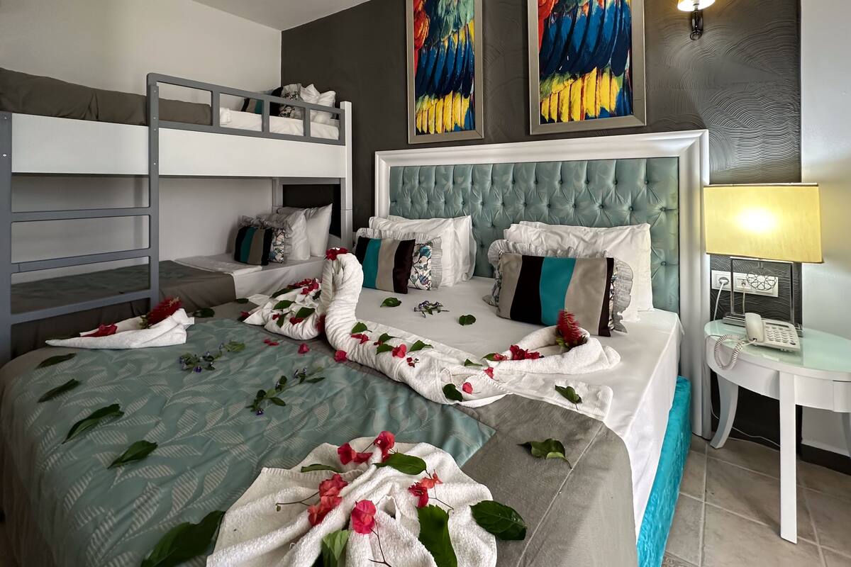 Quarto de hotel com uma cama de solteiro, uma cama grande com detalhes florais, uma mesinha de cabeceira com abajur e obras de arte nas paredes.