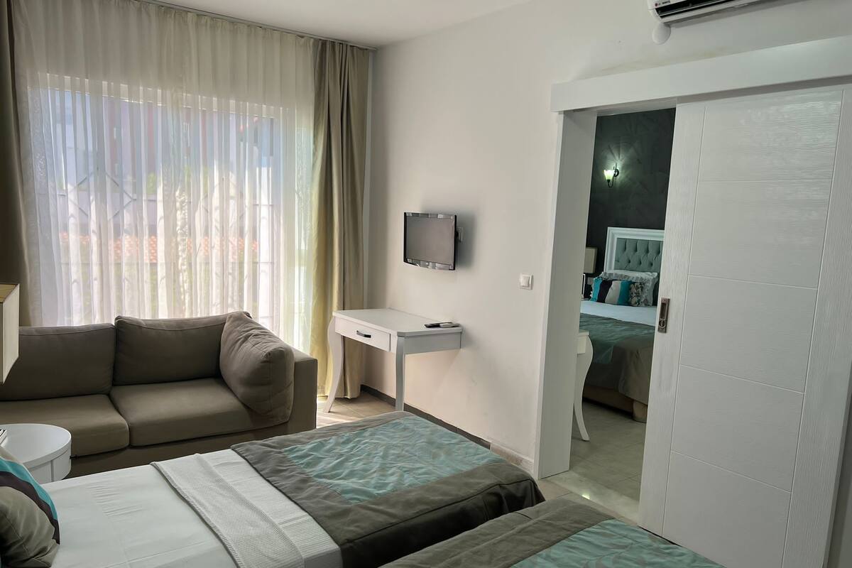 Quarto de hotel com sofá, duas camas de solteiro, escrivaninha, televisão e guarda-roupa.