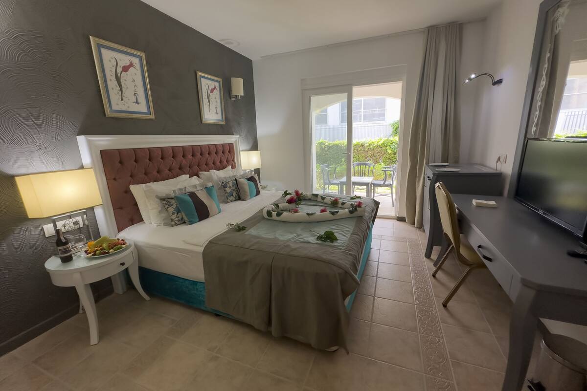 Quarto de hotel com cama, mesa de cabeceira, televisão e varanda com mobiliário exterior.
